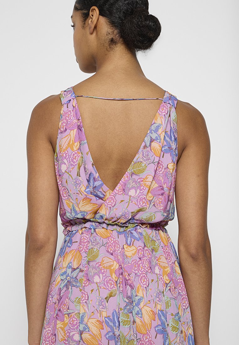 Kobieta z ciemnymi włosami spiętymi w kok, ubrana w bezrękawową sukienkę w kwiaty z głębokim dekoltem z tyłu i cienkim paskiem na górnej części pleców.