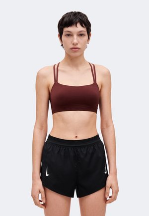 Une femme se tient face à l’avant sur un fond clair et uni. Elle porte un soutien-gorge de sport bordeaux et un short de sport noir.