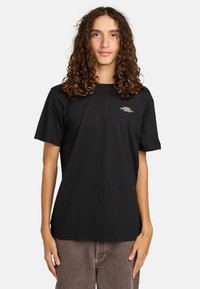 T-shirt in cotone nero con maniche corte; presenta un piccolo logo bianco sul petto. Il fit è rilassato e il tessuto appare morbido.