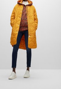 Abrigo largo acolchado amarillo con capucha, blusa roja y de estampado por debajo, jeans azul oscuro y zapatillas blancas. Presenta botones a presión y cremallera.