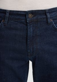 Jean en denim bleu foncé au niveau de la taille et de la cuisse supérieure, avec fermeture à bouton et détails de couture visibles.