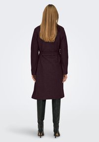 Femme aux longs cheveux blonds portant un manteau violet foncé ceinturé et des bottes noires montantes à talons, se tenant de dos devant un fond uni.