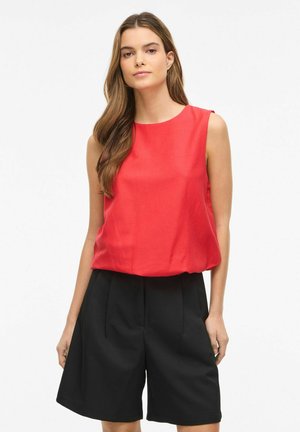 Kobieta z długimi brązowymi włosami, ubrana w czerwoną koszulę bez rękawów i czarne szorty z wysokim stanem, stojąca na jednolitym białym tle.