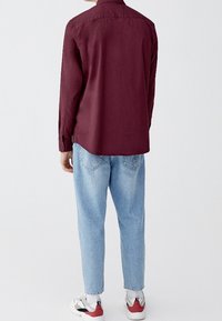 Bordeaux langemouwen shirt met een button-down kraag, gecombineerd met lichtblauwe jeans en witte sneakers met rode en zwarte accenten.