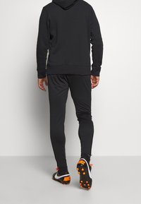Černá atletická mikina a kalhoty vyrobené z měkké tkaniny. Skládají se z délky po kotníky, žebrovaných manžet a zvýrazněných Nike obuvi s oranžovými detaily.
