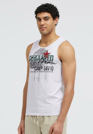 Man with curly dark hair wearing white sleeveless shirt with palm tree and text design, standing against plain light background.