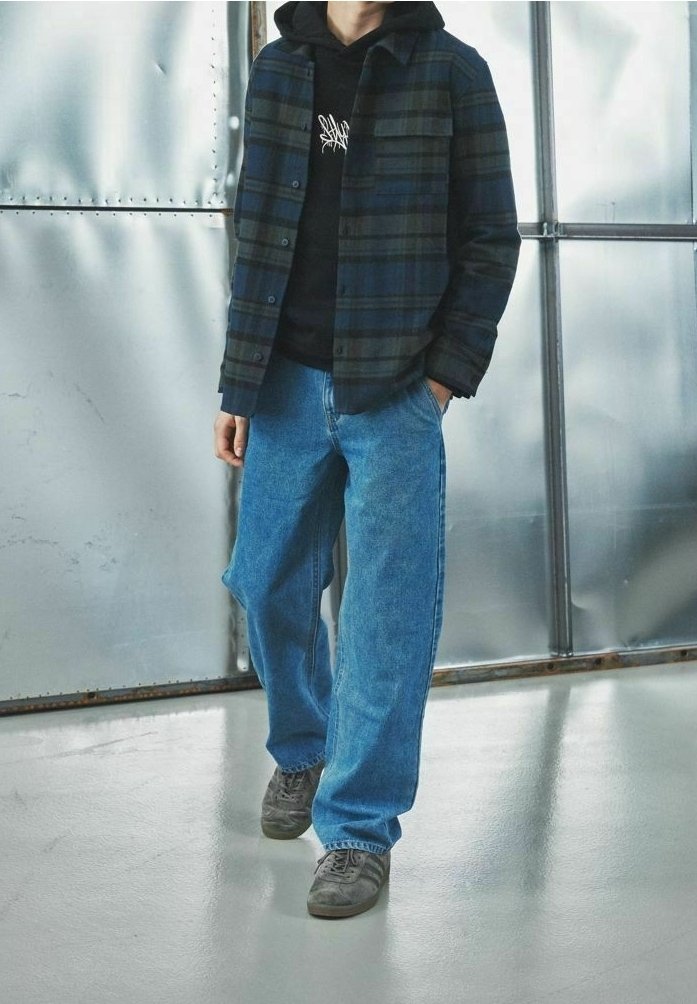 Niebieska koszula w kratkę z flaneli na czarnym hoodie, w komplecie z luźnymi niebieskimi dżinsami i szarymi sneakersami. Tło metalowe.