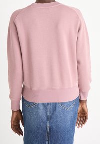 Różowy sweter z prążkowanymi mankietami i dołem, luźny krój, z rękawami raglanowymi. Noszony na dżinsowej spódnicy, pokazujący casualowy strój.