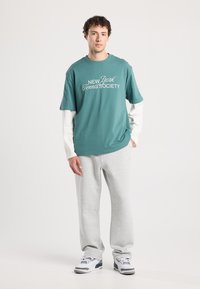 Tee-paita, jossa on teal-väri ja valkoinen käsiala "New York Tennis Society", kerroksittain pitkän valkoisen hihaisen paidan päällä, yhdistettynä harmaisiin verkkareihin ja lenkkareihin.