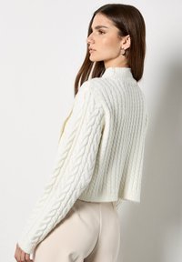 Maglione bianco lavorato a maglia con collo alto, caratterizzato da un design a coste e dettagli a trecce sulle maniche. Lunghezza corta con maniche lunghe.