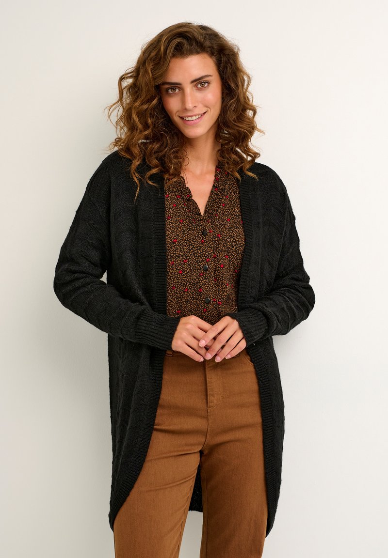 Zwarte gebreide cardigan met een open voorkant, gestructureerd patroon, lange mouwen, gecombineerd met een bruine blouse met patroon en bruine broek.