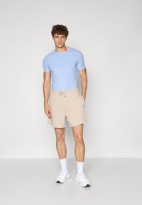 Helles, enges, kurzärmeliges Sportshirt in Blau, kombiniert mit beigen Shorts mit Kordelzug. Modell trägt weiße Sneaker und Knöchelsocken. Glatte Stofftextur.