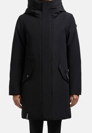 Manteau d'hiver long noir avec un col montant, une fermeture éclair frontale cachée et des poches à rabat à boutons-pression, présenté sur une personne devant un fond uni.