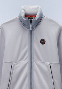 Hellgraue Zip-Jacke mit hohem Kragen, schwarzen Akzent-Reißverschlüssen und einem runden Logo auf der linken Brust. Hergestellt aus weichem, strukturiertem Stoff.