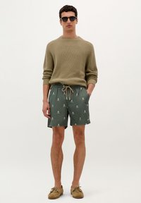 Costume de baie verzi cu model, cu un talie cu șnur, asortate cu un pulover tricotat bej. Modelul poartă ochelari de soare negri și pantofi loafers maro.