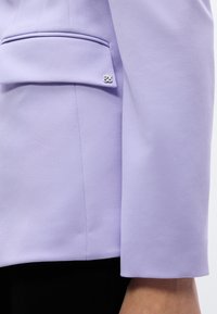 Κοντινό πλάνο στο μανίκι και την πτέρυγα της τσέπης ενός μωβ σακακιού με μεταλλικό σήμα "HUGO" στην πτέρυγα της τσέπης πάνω από μαύρο παντελόνι.