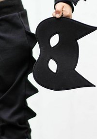 Svart tygmask formad som bokstaven 'B', med två ögonöppningar och en spetsig topp, hålls av en hand bredvid svart textilklädsel.