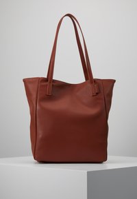 Bruine, textuurvolle tote bag met twee lange hengsels, open ontwerp en een zachte, flexibele vorm. Minimalistische stijl op een lichtgrijze achtergrond.