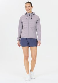 Smilende kvinde står iført en lys lilla hættet jakke, navy blå sportsshorts og hvide løbesko på en hvid baggrund.