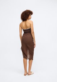 Brun strapless kjole i struktureret stof med en indsnævret talje og dekorative frynser i kanten, parret med simple sandaler.