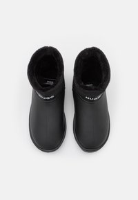 Czarne buty slip-on z miękkim futrzanym wykończeniem i logo "HUGO" na tylnej kołnierzu, widok z góry na białym tle.