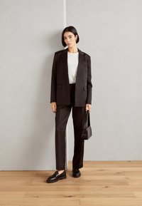 Black tailored suit with a single-button blazer and straight-leg trousers, paired with a white top and black loafers, holding a small black bag.