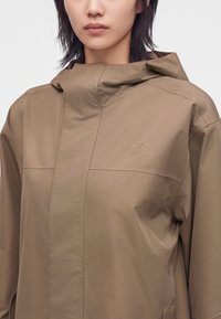 Jeune personne portant une veste de pluie à capuche marron clair avec une fermeture éclair dissimulée à l'avant et un logo de marque subtil sur la poitrine.