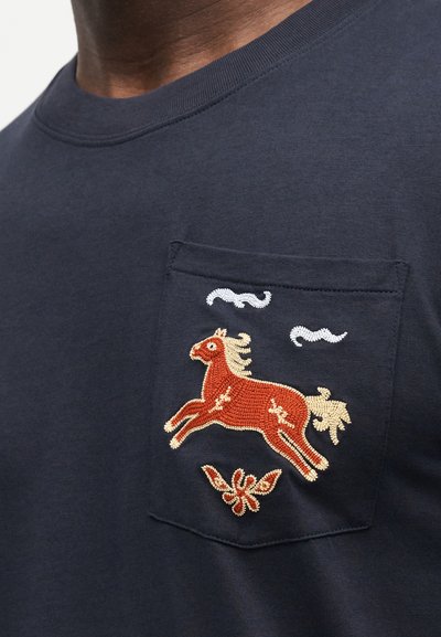 Τσέπη πουκαμίσου σε ναυτικό μπλε χρώμα με κεντημένο κόκκινο άλογο, εκρού χαίτη και ουρά, δύο λευκές νεφελώδεις μορφές και μικρό κόκκινο λουλουδένιο σχέδιο κάτω από το άλογο.