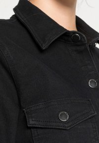 Mustat farkkutakki, jossa on terävä kaulus, napsautusnappit ja rintatasku, jossa on metalliosat ja yksityiskohtaista ommelta.