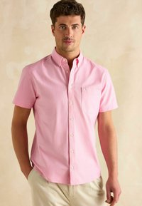 Svetloružová krátkopupočná košeľa s golierom na gombíky, prednou vreckom a bielymi gombíkmi, vyrobená z jemnej tkaniny s decentnou textúrou.