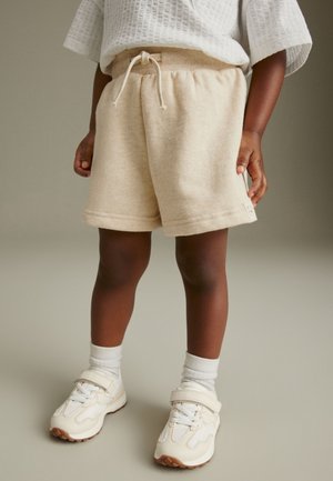 Barn iført beige shorts med snøre, hvide sokker og hvide sneakers med velcrolukning, stående på et neutralt gulv.