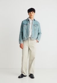 Νέος άνδρας που φοράει ανοιχτό μπλε τζιν μπουφάν, λευκό πουκάμισο, μπεζ παντελόνι και μαύρα παπούτσια, στέκεται μπροστά σε ένα απλό λευκό φόντο.