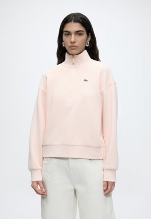 Frau trägt ein hellrosa Sweatshirt mit Viertelreißverschluss und kleinem grünem Krokodillogo sowie weiße Hosen, steht vor einem schlichten weißen Hintergrund.