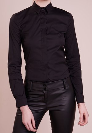 Person wearing a fitted black button-up shirt and black leather pants, standing against a plain beige background.