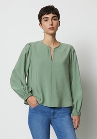 Blusa verde con una texture morbida e liscia, scollatura rotonda e dettaglio dei bottoni. Presenta maniche lunghe e arricciate e una vestibilità comoda.