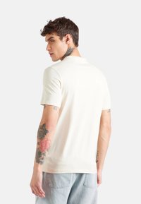 T-shirt corto color beige chiaro realizzato in tessuto morbido, con collo rotondo e vestibilità rilassata, indossato con jeans in denim blu chiaro.