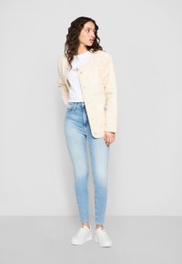 Jauna moteris su garbanotais plaukais, vilkinti bežinės spalvos čiužinį, baltą marškinėlį su logotipu, šviesiai mėlynas liesas džinsus ir baltus sportbačius, žiūri į dešinę.