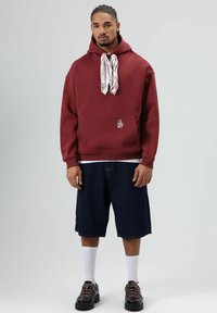 Bordeauxrode hoodie met een voorvak en wit logo, gecombineerd met donkere spijkershorts. Witte sokken en zwarte schoenen met roze veters maken de look compleet.