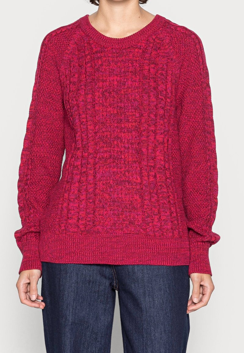 Personne portant un pull à manches longues en maille torsadée rose foncé et un jean bleu foncé, debout devant un fond clair uni.