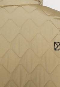 Beżowa pikowana tkanina kurtki z falistym wzorem i kontrastującym przeszyciem. Po prawej stronie znajduje się czarne logo.