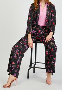 Femme assise sur un tabouret noir, portant un blazer et un pantalon noirs à fleurs roses et rouges, un haut côtelé rose et des sandales à talons hauts roses.