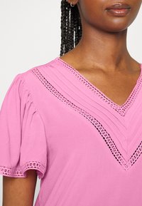 Blusa rosa com mangas curtas bufantes, apresentando costura detalhada e um decote em V, complementada por acabamentos em renda ao longo do decote.