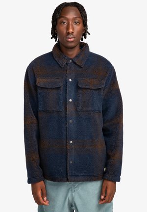 Veste en polaire bleu marine avec un motif en tartan marron, deux poches poitrine, fermeture à boutons et col. Matière douce et texturée.
