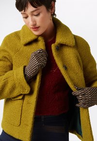 Veste en mélange de laine jaune avec une finition texturée, dotée d'un large col, de deux boutons et d'une poche latérale, portée par-dessus un pull rouge profond.