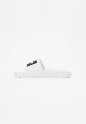White slide sandals with a black logo strap. Made of smooth plastic, featuring a flat sole with ridged detailing for grip.