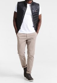 Uomo che indossa un gilet trapuntato nero, una maglietta bianca, pantaloni beige e sneakers marroni scuri, che cammina con le mani in tasca su uno sfondo bianco.