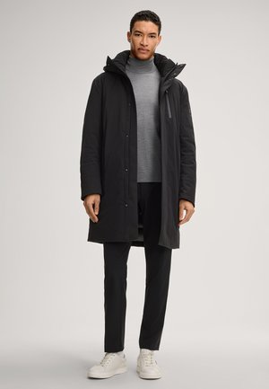 Czarna ocieplana parka z odpinanym kapturem, zapinana na zamek i zatrzask, zestawiona z szarym golfem i czarnymi spodniami, noszona z białymi sneakersami.