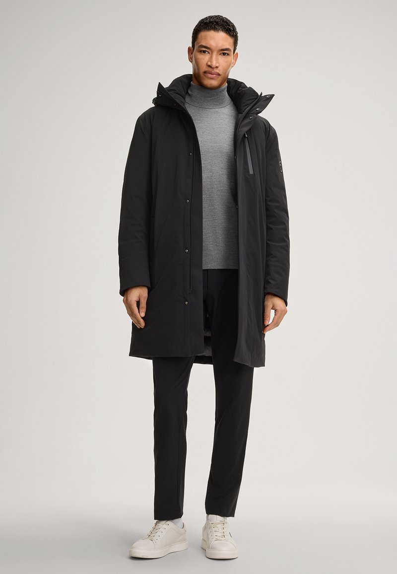 Czarna ocieplana parka z odpinanym kapturem, zapinana na zamek i zatrzask, zestawiona z szarym golfem i czarnymi spodniami, noszona z białymi sneakersami.