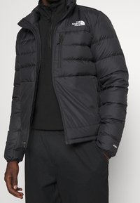 Man wearing black puffer jacket with "The North Face" logo on chest and zippered chest pocket, with hands in black pants pockets.