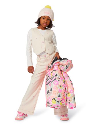 Gilet cropped beige avec des motifs de cœurs roses, associé à un pantalon taille haute rose clair. Le mannequin tient une veste en duvet colorée avec un design abstrait.